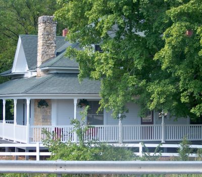 A quaint white house with a large stone chimney and a spacious wraparound porch. The house has a green roof and is nestled among lush green trees. A metal guardrail runs along the bottom of the image.