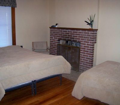 A simple bedroom with light beige walls and hardwood floors features a brick fireplace. There are two beds, both with beige bedspreads, and a small chair near the fireplace. A small plant and bird figurine adorn the mantle. White blinds cover the window.