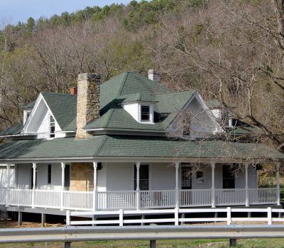 A white farmhouse style house with a wraparound porch. There is a stone chimney and the roof of the house is a light green. It is in the foreground, and a wooded mountain is in the background.