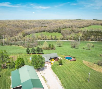 Aerial view of a rural property with a green-roofed building, a house, trees, a round pool, and a pond, surrounded by expansive grassy fields and forested hills under a blue sky.
