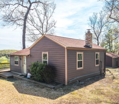 A small brown wooden house with a red roof sits among trees, with a gravel driveway and a deck overlooking a green, hilly landscape under a blue sky.