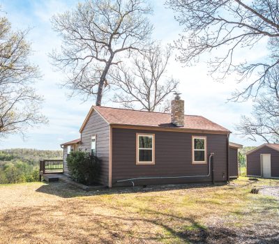 A small brown cabin with a red roof sits among leafless trees on a sunny day, with a gravel driveway, deck, and a small shed nearby. Rolling hills and greenery are visible in the background.