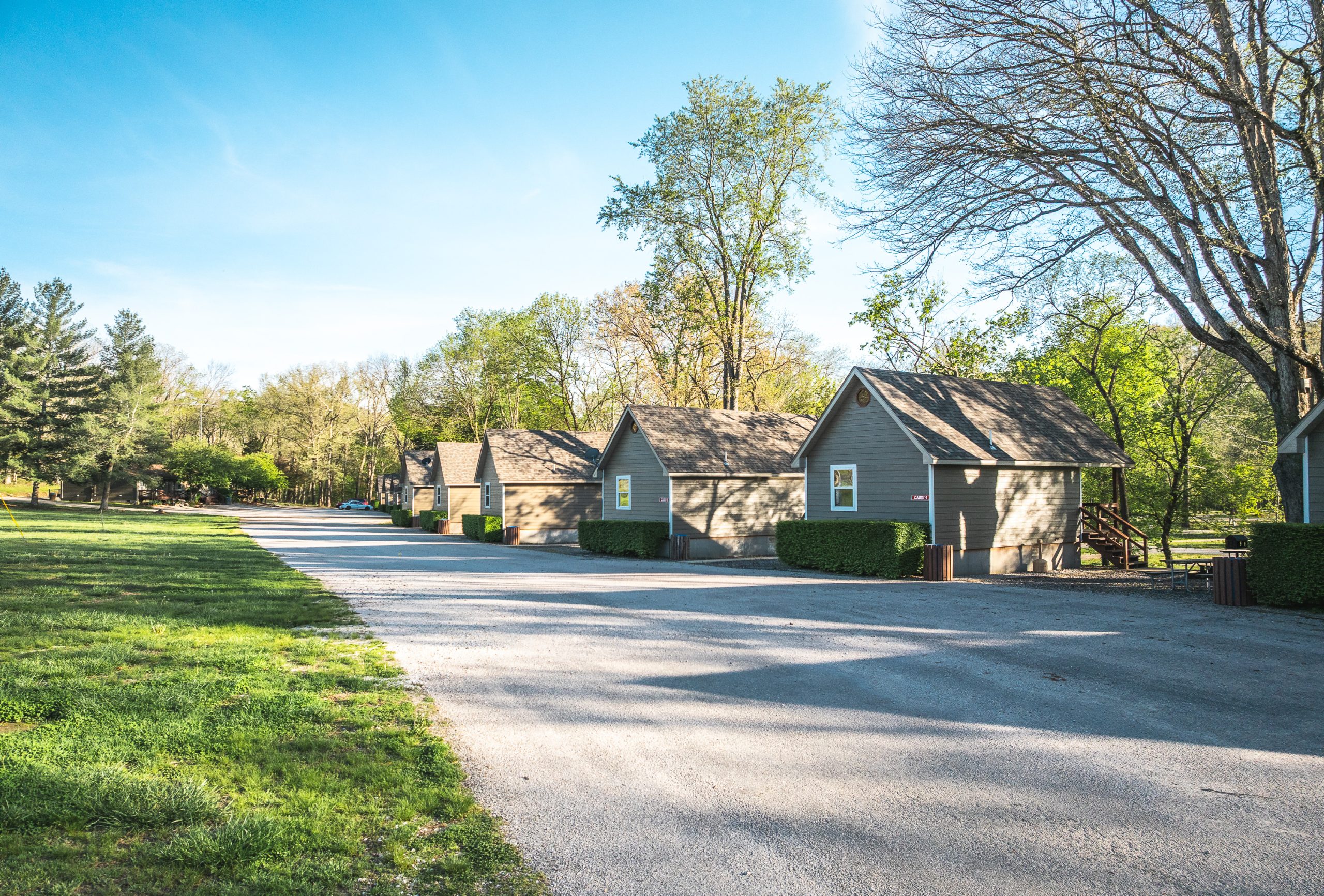 A row of small, gray cabins with peaked roofs lines a sunlit gravel road, surrounded by green grass and trees under a clear blue sky.