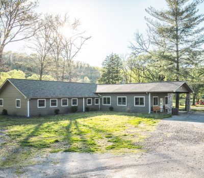 A single-story, light green house with a large front porch sits surrounded by trees and grassy areas under bright sunlight, with a gravel driveway in front and a wooded hillside in the background.