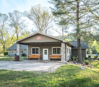 A small, rustic building with stone pillars and wooden benches on a porch, surrounded by trees and grass under a clear sky. A red sign is mounted above the entrance door.