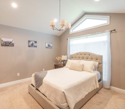 A cozy bedroom with neutral walls, a tufted bed with cream bedding, a wooden nightstand, a lamp, and three black-and-white animal prints on the wall. A large window lets in natural light.