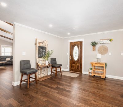 Bright, spacious entryway with wood floors, a wooden front door, two gray chairs, a chalkboard on an easel, and a small rolling cart. Living room visible on the left, hallway on the right.
