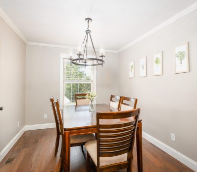 A bright dining room with a wooden table and six matching chairs, a modern chandelier overhead, light-colored walls, hardwood floors, a window with sunlight, and framed artwork on the wall.