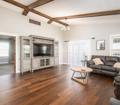 A spacious living room with wood floors, a dark sectional sofa, a round wooden coffee table, a large TV in a light wood entertainment center, ceiling beams, and windows letting in natural light.