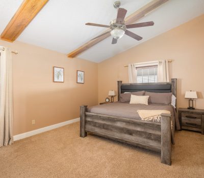 A bedroom with beige walls and carpet, a wooden bed with gray bedding, two nightstands with lamps, two wall art pieces, two windows with blinds, and a ceiling fan with wooden beams visible on the ceiling.