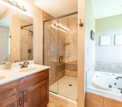Bright bathroom with a double-sink vanity, glass-enclosed shower with beige tiles, and a corner bathtub with bronze fixtures, next to a wall with decorative frames.