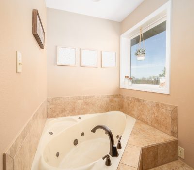 A beige-tiled bathroom features a corner jetted bathtub with a dark faucet, a large window with a hanging plant, framed art on the wall, and natural light filling the space.