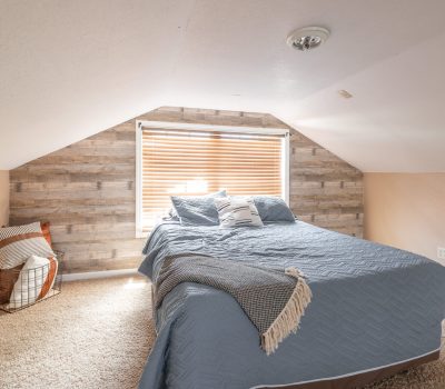 Cozy attic bedroom with sloped ceiling, a large bed covered in blue bedding, a basket with pillows, a window with blinds, and wood-paneled accent wall. Soft beige carpet covers the floor.