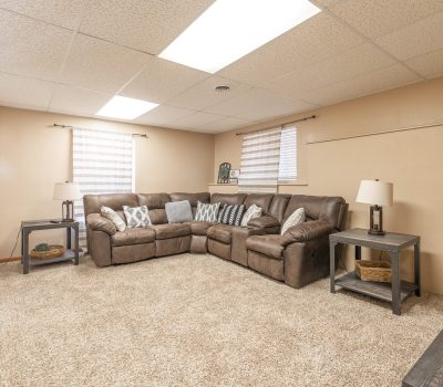 A cozy basement living room with beige walls, carpeted floor, brown sectional sofa with patterned pillows, two end tables with lamps, and two windows with blinds letting in natural light.
