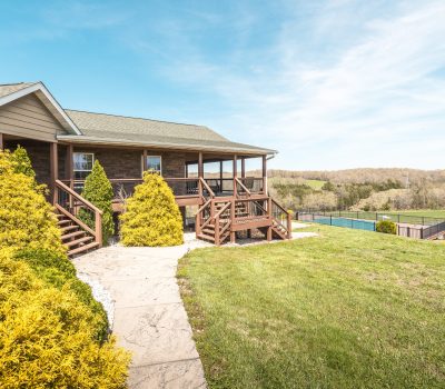 A wooden house with a covered porch sits on a grassy hill, surrounded by bushes. A stone path leads to the steps. In the background, there is a pond, fencing, and rolling hills under a blue sky.