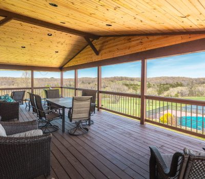 A spacious covered wooden deck with patio furniture overlooks a green landscape and a blue swimming pool on a sunny day. The ceiling is wood-paneled and the railing offers clear views of the fields and distant trees.