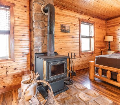 A cozy log cabin room with wood-paneled walls and ceiling, featuring a stone hearth with a black wood-burning stove, firewood in a basket, and a rustic wooden bed in the background.