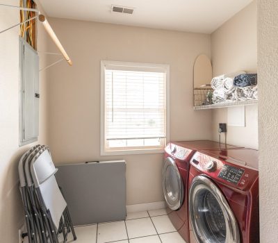 A small laundry room with a window, red washer and dryer, folding chairs, a wall-mounted drying rod, a shelf with towels, and a tile floor. Natural light comes in through the window.