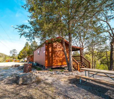 A small wooden cabin with a front porch sits among tall trees, next to a gravel area with a picnic table and a grill, under a clear blue sky in a rural setting.