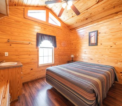A cozy bedroom with wood-paneled walls and ceiling, a large window with a dark curtain, a double bed with a striped quilt, a small desk with a sink, and a ceiling fan with lights.