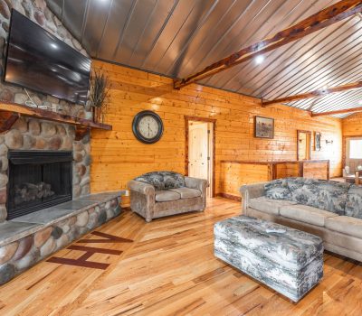 A cozy living room with wood-paneled walls, a stone fireplace, a mounted TV, plush sofas, an ottoman, hardwood floors, and exposed beams on a metal ceiling.