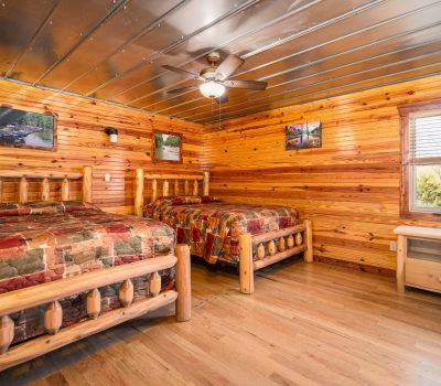 A cozy cabin bedroom with two rustic wooden beds, colorful patchwork quilts, wood-paneled walls and floor, a window with blinds, a small wooden table, ceiling fan, and landscape photos on the walls.