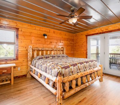 A cozy log cabin bedroom with wood-paneled walls and floors, a rustic wooden bed with a patchwork quilt, a matching nightstand, and a ceiling fan. French doors open to a porch with outdoor seating.
