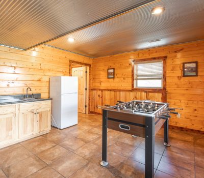 A rustic game room with wood-paneled walls, tiled floor, a foosball table in the center, a white refrigerator, a counter with a sink, and windows letting in natural light.