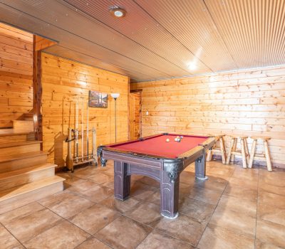 A cozy wooden game room with a red pool table, cues on a rack, wooden barstools, tiled floor, and a staircase with wooden steps leading upward. A framed picture and a floor lamp are on the back wall.