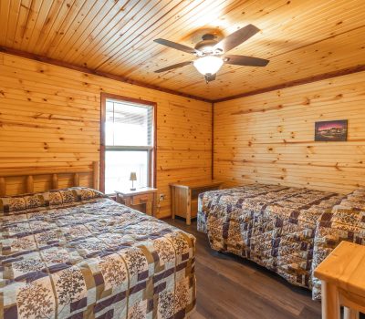 A cozy cabin bedroom with wood-paneled walls and ceiling, two double beds with patchwork quilts, a ceiling fan with a light, a window, a small nightstand, and a landscape picture on the wall.
