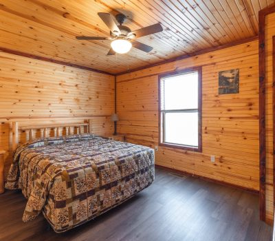 Cozy bedroom with wood-paneled walls and ceiling, a bed with a patchwork quilt, a wooden nightstand, window with natural light, ceiling fan, and a wall-mounted TV.