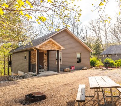 A small brown cabin with a front porch sits among trees on a sunny day. A picnic table and fire pit are in the foreground, and a "For Sale" sign is posted near the cabin. Another cabin is visible in the background.