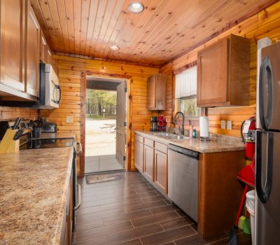 A cozy cabin kitchen with wooden walls and ceiling, brown cabinets, stainless steel appliances, and granite countertops. A door at the end opens to an outdoor picnic area with trees and a picnic table visible.