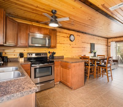 A cozy kitchen with wood-paneled walls and ceiling, stainless steel appliances, brown cabinets, granite countertops, a ceiling fan, and an adjacent dining area with a table and chairs by large windows.