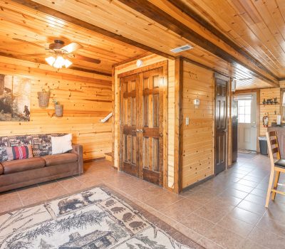 A cozy cabin interior with wooden walls and ceiling, a brown couch with pillows, a nature-themed rug, a ceiling fan, and a small dining area next to a kitchen. Warm lighting enhances the rustic decor.