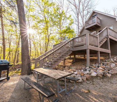 A wooden cabin with an elevated deck sits among trees and rocks. A picnic table and a barbecue grill are nearby, with sunlight streaming through the forest in the background.