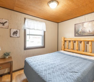 A cozy bedroom with a rustic wooden bed frame, blue quilt, bedside table with a lamp and plant, window with white curtains, and nature-themed wall art and decorations.