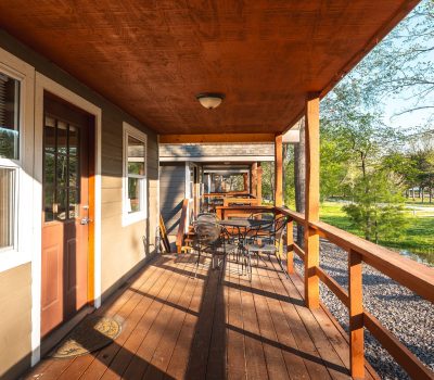 Sunny wooden porch with a table and chairs, overlooking a grassy area with trees. The porch is attached to a tan house with white-trimmed windows and a brown door. Shadows stretch across the wooden floor.