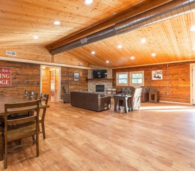 A spacious lodge room with wood-paneled walls and ceiling, featuring a dining table and chairs, a cozy seating area with sofas, a TV, a fireplace, and large windows letting in natural light. A sign reads "Huzzah Bob's Dining Lodge.