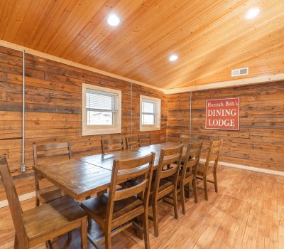 A rustic dining room with wood-paneled walls and ceiling, a long wooden table with ten chairs, two windows, and a sign on the wall reading “Huzzah Bob’s Dining Lodge.” Bright overhead lighting.