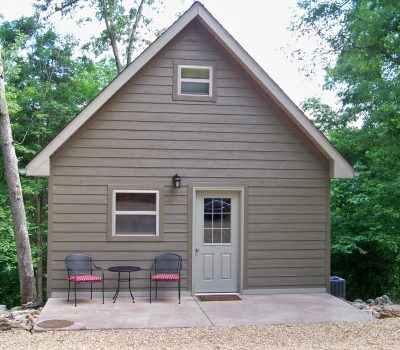 A small, gray wooden house with a steeply pitched roof sits among trees. The house has a single front door, two windows, and a small concrete patio with two black metal chairs and a round table, both with red and white striped cushions.
