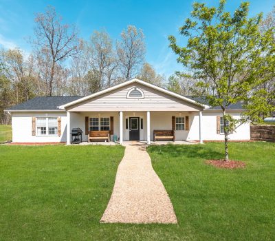 Single-story white house with a gabled roof, two benches on the front porch, a grill, a tree in the yard, and a gravel path leading to the entrance. The house is surrounded by grass and trees under a blue sky.