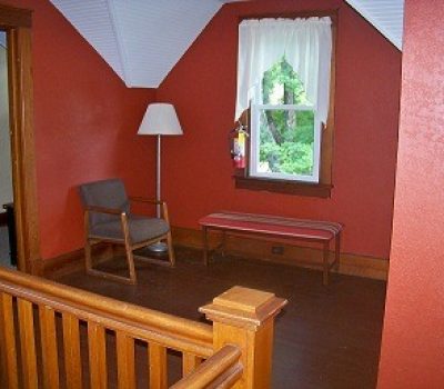 A cozy room with sloped ceilings painted in a warm red tone. There is a window with white curtains, a floor lamp, a gray cushioned armchair, and a red-striped bench. The foreground features a wooden staircase railing. Natural light filters through the window.