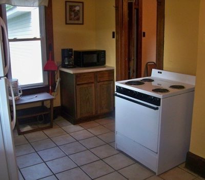 A small kitchen with a white stove in the center, brown wooden cabinets, a counter with a microwave, and a window with natural light. The walls are yellow, and there is a picture frame and a red lamp on the countertop. The floor is tiled with light-colored squares.
